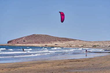 İspanya, Kanarya Adaları, Tenerife, Playa del Medano - 05 Haziran 2025: Kadın arka planda tepelerle kıyı şeridi boyunca yürürken erkek uçurtmalar