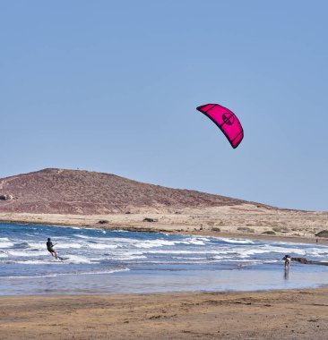 İspanya, Kanarya Adaları, Tenerife, Playa del Medano - 05 Haziran 2025: Kadın arka planda tepelerle kıyı şeridi boyunca yürürken erkek uçurtmalar
