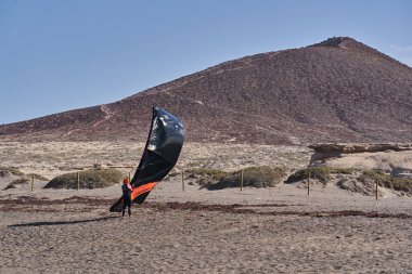 İspanya, Kanarya Adaları, Tenerife, Playa del Medano - 05 Haziran 2025: Kitesurfer arka planda Montana Roja volkanik tepesi ile kumsalda siyah bir tepe hazırlıyor