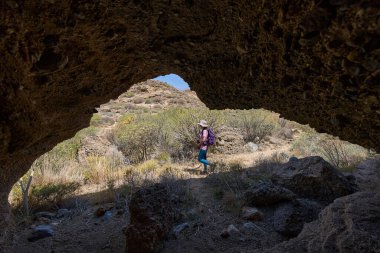 Kuru bir kanyon manzarasında volkanik bir mağaranın girişinden geçen kadın yürüyüşçü.