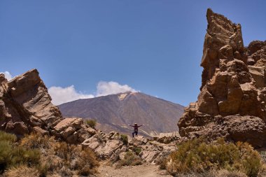 Kolları açık kayaların üzerinde duran kadın yürüyüşçü, arkasında Pico del Teide ile Roques de Garcia tarafından çerçevelenmiş.
