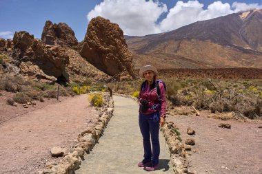 Kameralı ve sırt çantalı bir bayan yürüyüşçü. Arka planda volkanik oluşumlar ve Teide volkanı olan bir patikada duruyor.