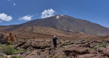 Volkanik pahoe arazisinde duran bir adam açık mavi gökyüzünün altında Pico del Teide 'yi fotoğraflıyor.