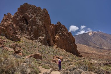 Roques de Garcia 'da açık ve güneşli bir günde sırt çantası ve kamerasıyla kayalık bir volkanik yolda yürüyen kadın yürüyüşçü ve seyahat fotoğrafçısı.