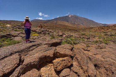 Dişi yürüyüşçü, Teide stratovolcano ile açık yaz gökyüzünün altında engebeli bir lav tarlasında manzara fotoğrafı çekiyor.