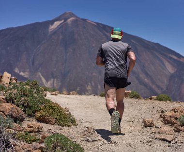 Pico del Teide 'nin arka planında yükselen güneşli bir volkanik yamaçta Guajara Dağı' na tırmanan bir patika koşucusu.