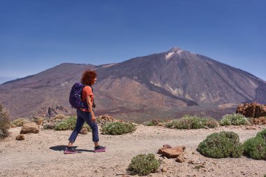 Pico del Teide volkanı ve arka planda engebeli volkanik arazisi olan tozlu bir patikada yürüyen kadın yürüyüşçü.