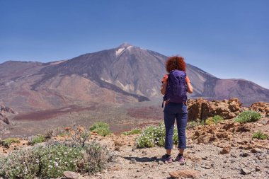 Uçsuz bucaksız volkanik manzaraya ve Teide Ulusal Parkı 'ndaki Pico del Teide' e bakan kayalık yolda duran kadın yürüyüşçü.