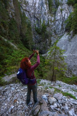 Kıvırcık kızıl saçlı ve sırt çantalı yürüyüşçü kadın, Romanya 'nın Apuseni Dağları, Cetatile Ponorului' de kayalık bir patikada telefonuyla uçurumları fotoğraflıyor.