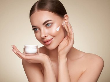 Güzellik kraliçesi yüzüne krem sürüyor. Temiz tenli genç bir kadın. Yüz kremi kavanozu olan bir model.