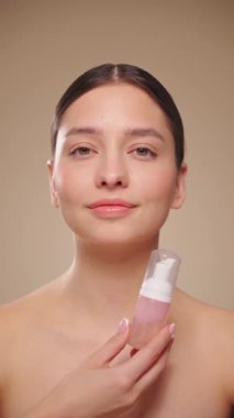 Genç bir kadın yıkamak için köpüklü bir şişeyle hava atıyor. Günlük cilt bakımı kozmetik reklamı. Stüdyo çekimi bej arka plan. Yavaş çekim, dikey video