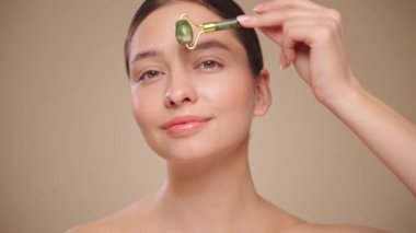 Doğal bir yeşim taşlı rulo kullanırken rahat ve gülümseyen genç bir kadının videosunu kapat. Deri silindirli yaşlanma önleyici ve kozmetik temizlik için yüz bakımı. Yavaş çekim
