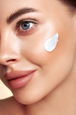 Güzel bir kadın yüzüne nemlendirici krem sürüyor. Bej arka planda mükemmel makyajlı gülümseyen bir kadının fotoğrafı. Güzellik konsepti