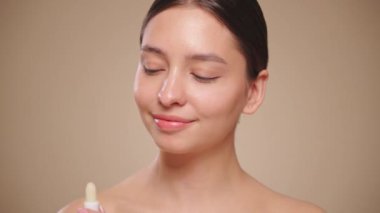 Genç bir kadın, bej arkaplandaki dudakları nemlendirmek için koruyucu ruj ya da merhem sürüyor. Dudak bakımı kozmetik reklam konsepti. Yavaş çekim