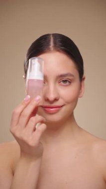 Genç bir kadın yıkamak için köpüklü bir şişeyle hava atıyor. Günlük cilt bakımı kozmetik reklamı. Stüdyo çekimi bej arka plan. Yavaş çekim, dikey video