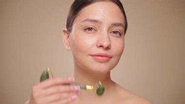Doğal bir yeşim taşlı rulo kullanırken rahat ve gülümseyen genç bir kadının videosunu kapat. Deri silindirli yaşlanma önleyici ve kozmetik temizlik için yüz bakımı. Yavaş çekim
