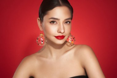 Kırmızı arka planda güzel bir genç kadının portresi, kırmızı ruj ve festival makyajı, Noel oyuncak küpeler, parlak cilt, kış tatili konsepti.