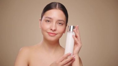 Gülen kadın elinde kozmetik sütle şişe tutuyor. Bej arka planda mükemmel makyajlı çekici bir kadın videosu. Güzellik konsepti. Yavaş çekim
