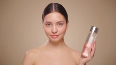 Gülen kadın elinde kozmetik tonikle şişeyi tutuyor. Bej arka planda mükemmel makyajlı çekici bir kadın videosu. Güzellik ve cilt bakımı. Makyaj kaldırılıyor. Yavaş çekim
