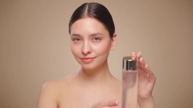 Gülen kadın elinde kozmetik tonikle şişeyi tutuyor. Bej arka planda mükemmel makyajlı çekici bir kadın videosu. Güzellik ve cilt bakımı. Makyaj kaldırılıyor. Yavaş çekim
