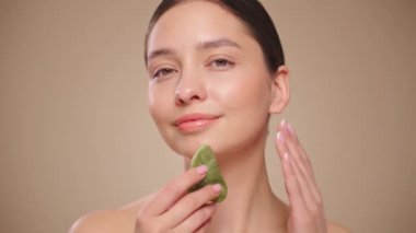 Cilt bakımı rutini yapan güzel genç bir kadın, hurda, yüz masajı, yeşim taşıyla yüz bakımı, yaşlanma ve kozmetik temizleme için yüz bakımı. Yavaş çekim