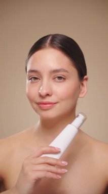 Bir şişe nemlendirici kremi olan güzel bir kadın. Bej arka planda mükemmel cilde sahip bir kadının videosu. Güzellik ve Deri Bakımı konsepti. Yavaş çekim, dikey video