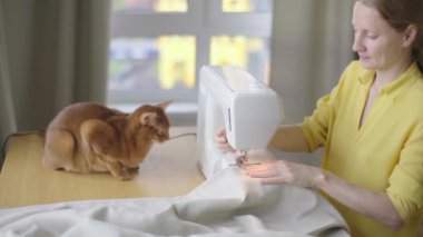 Dikiş makinesiyle elbise yapan genç bir kadın. Evdeki iş yerinde oturuyor.