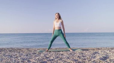 Genç kadın deniz kenarındaki plajda yoga yapıyor. Yoga egzersizleri yapan bir kız, ve deniz sörfünün arka planına ve güzel bir gökyüzüne karşı