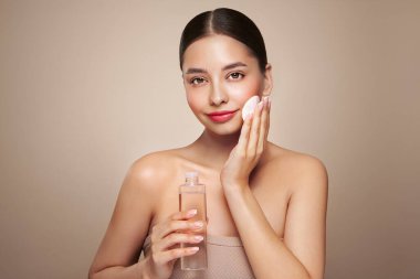 Gülen kadın elinde kozmetik tonikle şişeyi tutuyor. Bej arka planda mükemmel makyajlı çekici bir kadın fotoğrafı. Güzellik ve cilt bakımı. Makyaj kaldırılıyor