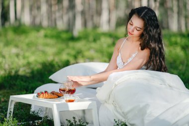 Kadın bir yaz ormanında uyanıyor. Genç bir kadın yaz ormanında yatakta kahvaltı yapıyor.