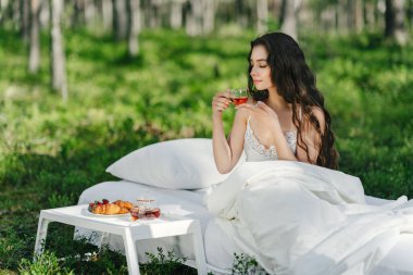 Kadın bir yaz ormanında uyanıyor. Genç bir kadın yaz ormanında yatakta kahvaltı yapıyor.