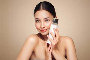 Gülen kadın elinde kozmetik sütle şişe tutuyor. Bej arka planda mükemmel makyajlı çekici bir kadın fotoğrafı. Güzellik konsepti