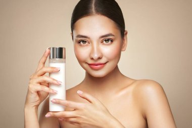 Gülen kadın elinde kozmetik sütle şişe tutuyor. Bej arka planda mükemmel makyajlı çekici bir kadın fotoğrafı. Güzellik konsepti