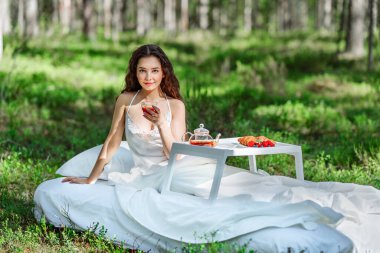 Kadın bir yaz ormanında uyanıyor. Genç bir kadın yaz ormanında yatakta kahvaltı yapıyor.