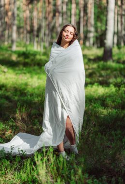 Sıcak bir battaniyeye sarılmış genç bir kadın yaz ormanında duruyor. Kız yaz ormanında uyanıyor. Eko-konfor ve rahatlama