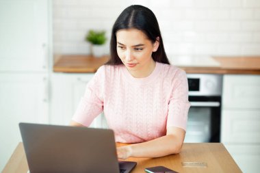 Ev mutfağında dizüstü bilgisayarla çalışan güzel bir genç kadın. Evde çalış. Serbest, uzak çalışma