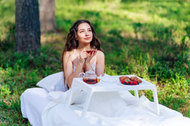 Kadın bir yaz ormanında uyanıyor. Genç bir kadın yaz ormanında yatakta kahvaltı yapıyor.