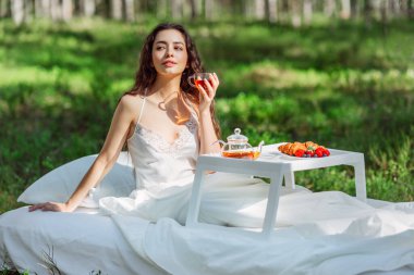 Kadın bir yaz ormanında uyanıyor. Genç bir kadın yaz ormanında yatakta kahvaltı yapıyor.