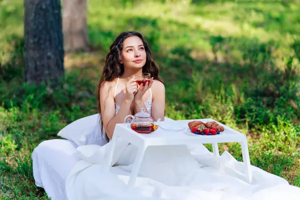 Kadın bir yaz ormanında uyanıyor. Genç bir kadın yaz ormanında yatakta kahvaltı yapıyor.