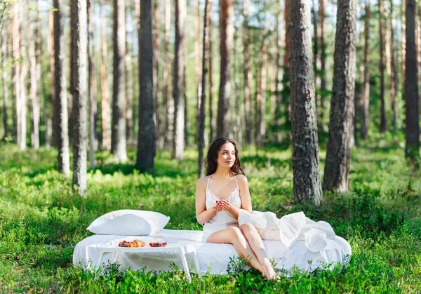 Kadın bir yaz ormanında uyanıyor. Genç bir kadın yaz ormanında yatakta kahvaltı yapıyor.