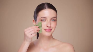 Cilt bakımı rutini yapan güzel genç bir kadın, hurda, yüz masajı, yeşim taşıyla yüz bakımı, yaşlanma ve kozmetik temizleme için yüz bakımı. Yavaş çekim