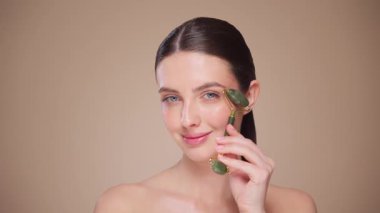 Doğal bir yeşim taşlı rulo kullanırken rahat ve gülümseyen genç bir kadının videosunu kapat. Deri silindirli yaşlanma önleyici ve kozmetik temizlik için yüz bakımı. Yavaş çekim