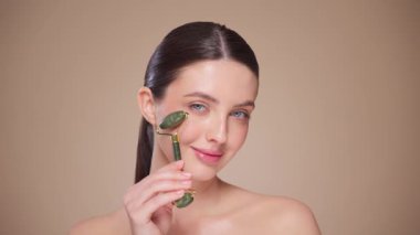 Doğal bir yeşim taşlı rulo kullanırken rahat ve gülümseyen genç bir kadının videosunu kapat. Deri silindirli yaşlanma önleyici ve kozmetik temizlik için yüz bakımı. Yavaş çekim