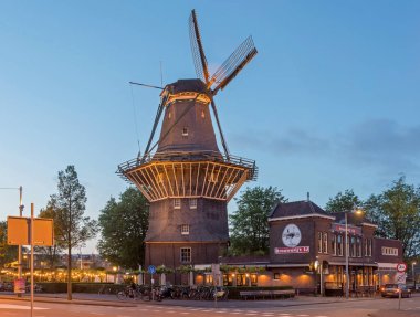 Amsterdam, Netherlands - May 16, 2018: Industrial Style Brewery Ostrich Next to a Traditional Windmill Gooyer Historic Landmark at Dusk. clipart