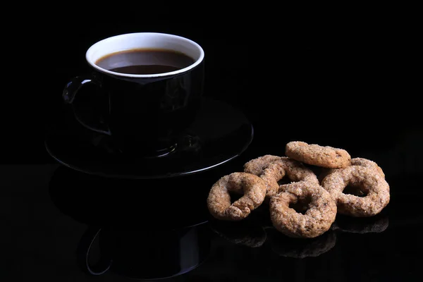 A cup of hot coffee and coffee rings biscuits on black.