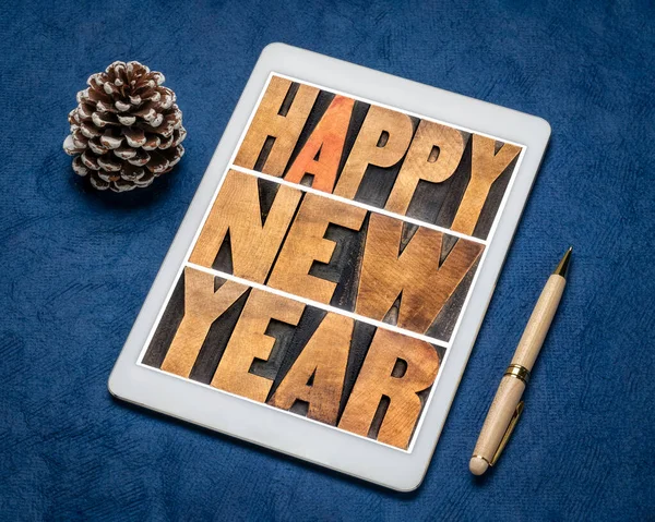 happy new year typography in vintage letterpress wood type on a digital tablet against blue handmade paper with a frosty pine cone, greeting card concept