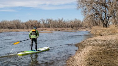 Erkek kürekçi, Colorado 'daki South Platte Nehri' nin ilk bahar manzarasında şişme bir kürek üstünde akıntıya karşı kürek çekiyor.