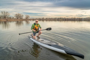 Kıdemli adam (Mid 60s) bir stand up ile Paddleboard bir gün batımı, Colorado erken bahar göl sahne onun kürek egzersiz bitirme.