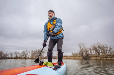 Yaşlı erkek raketçi soğuk kış havasında sakin bir gölde ayakta kürek çekiyor.