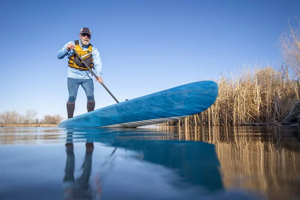 シニア男性パドラーは春に穏やかな湖の上にスタンドアップパドルボードを漕いでいます 水位でのアクションカメラからカエルの視点 — ストック写真